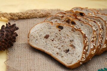 Several slices of multigrain bread rest on a wooden plate atop burlap fabric, surrounded by natural decor like pine cones and wheat.
