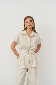 Woman with long brown hair wearing beige belted top and pants poses with hands on hips. She has large hoop earrings and a neutral expression. Light background, casual attire style