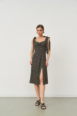 Woman in a knee-length sleeveless black dress with white dots and tied shoulder straps stands with...