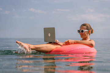 A woman is floating on a pink inflatable raft in the ocean while holding a laptop. Concept of relaxation and leisure, as the woman is enjoying her time in the water while working on her laptop.