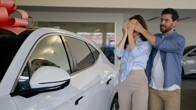 Caucasian lovely man male guy husband boyfriend make surprise present gift close eyes to woman female girl wife girlfriend amazed wonder happy couple buying new car hugging auto in showroom automobile