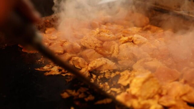 Marinated chicken pieces sizzle and cook on a hot flat top grill. A spatula turns the meat, creating steam. Perfect for delicious, hot, freshly prepared food.