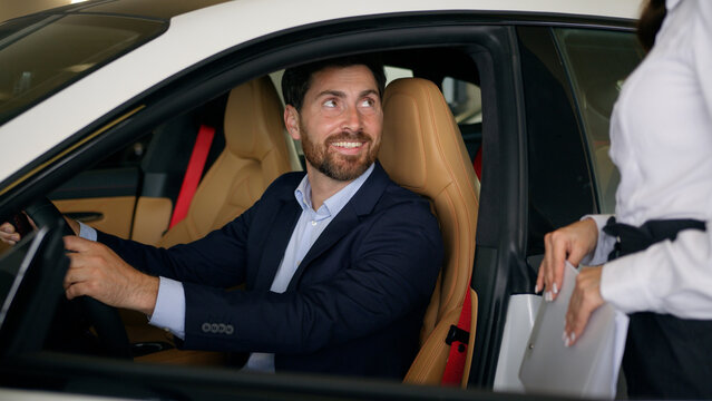 Caucasian woman dealer saleswoman female consultant girl help talking showing new luxury electric car to male client buyer man guy choose buying automobile auto sitting inside in salon rent service