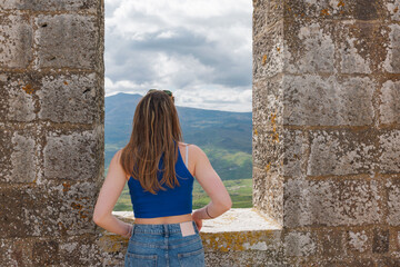 A young woman gazes at the breathtaking landscape of Tuscany from a stone window. The rolling hills...
