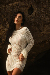 A slim brunette model in a short white crochet dress stands in shallow sea water, posing sensually...