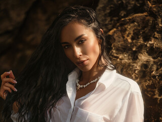 Young brunette woman in a soaked white shirt kneels near a rocky shore, posing seductively under...