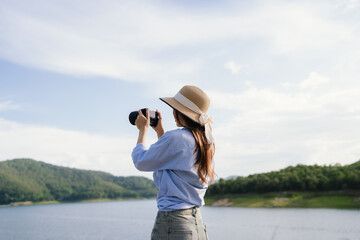 A young Asian woman leans on a white car, gazing thoughtfully at a serene mountain lake, embracing...