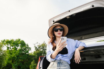 A young Asian woman leans on a white car, gazing thoughtfully at a serene mountain lake, embracing...