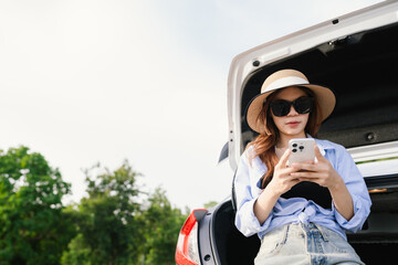 A young Asian woman leans on a white car, gazing thoughtfully at a serene mountain lake, embracing...