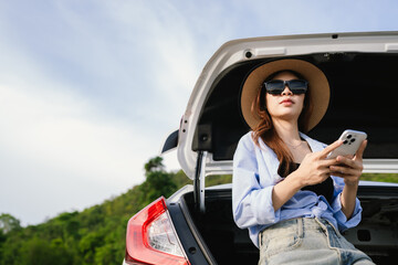 A young Asian woman leans on a white car, gazing thoughtfully at a serene mountain lake, embracing...