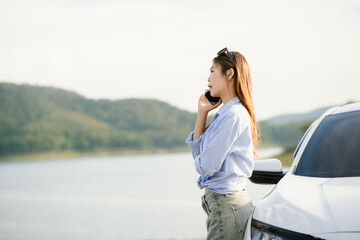 A young Asian woman leans on a white car, gazing thoughtfully at a serene mountain lake, embracing...