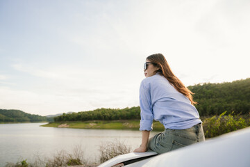 A young Asian woman leans on a white car, gazing thoughtfully at a serene mountain lake, embracing...
