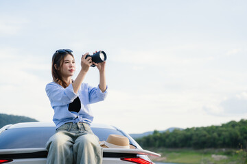 A young Asian woman leans on a white car, gazing thoughtfully at a serene mountain lake, embracing...
