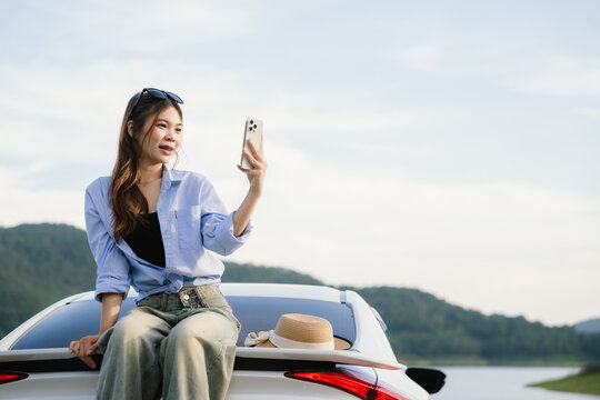 A young Asian woman leans on a white car, gazing thoughtfully at a serene mountain lake, embracing peace, nature, freedom, and the spirit of exploration.
