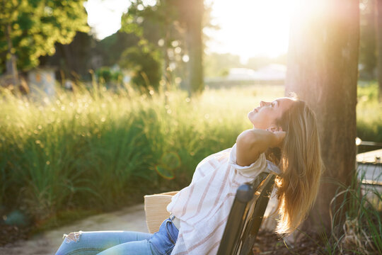 Woman relaxing outdoors on bench. Mindfulness, slow living, wellness, self-care in sunny summer park. Green escape, connecting with nature. Urban rewilding, climate resilience. Sustainable lifestyle - Powered by Adobe