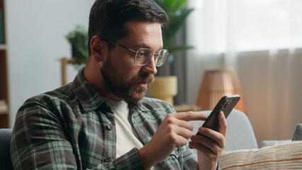 Obraz na płótnie Canvas Caucasian man male guy user at home browsing mobile phone app social media smartphone shock bad news amazed trouble problem confused puzzled wonder online internet web at home couch sofa apartment