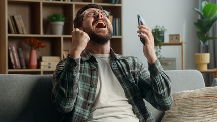 Cheerful rejoice happy winner champion yelling happiness man at home guy male gamer user using mobile phone shout joy win video game online achieve victory winning smartphone at home couch celebrate