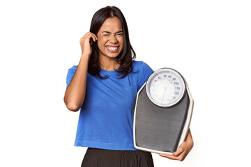 Filipino woman with scale in studio covering ears with hands.