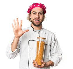 Young chef holds pasta sauce jar in studio smiling cheerful showing number five with fingers.
