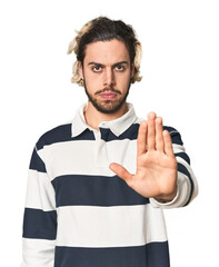 Young Caucasian man in studio setting standing with outstretched hand showing stop sign, preventing...