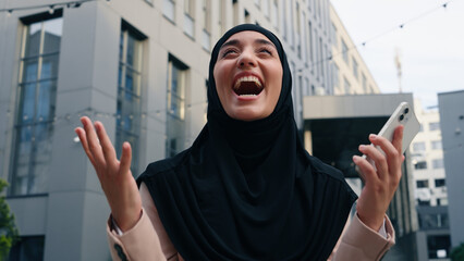 Happy excited winner Muslim business woman Islamic businesswoman girl female in black hijab employer rejoice good news mobile phone success winning victory smartphone in city yelling company achieve