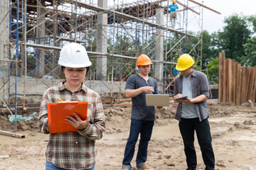 Asian female engineer writing on clipboard while two male workers review plans on digital devices at a construction site. Teamwork and supervision in civil engineering project.