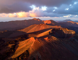 Aerial view of volcanic cones and craters in a mountainous landscape under a dramatic sky at sunset.