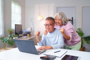 Fototapeta premium Happy Older Asian Couple Celebrating Success at Home on Laptop. Senior Couple Enjoying Retirement. Home Office Setup for Elderly with Modern Tech