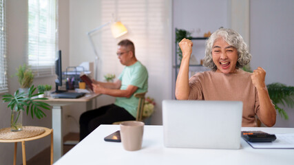 Obraz premium Senior Asian Women Celebrate Success with Laptop At Home While Husband Reads in Background Happy Retirement Lifestyle Well Being Concept