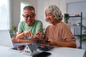 Senior Asian Couple Planning Retirement Finances at Home with Laptop, Calculator and Model House...