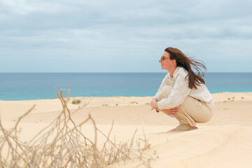 Obraz premium A woman sits thoughtfully on a sandy beach, gazing towards the ocean, with wind in her hair and a calm expression on her face.