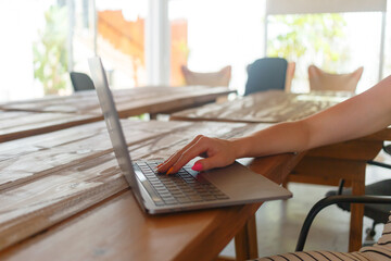 A person types on a laptop at a spacious wooden table in a cafe with natural light illuminating the...