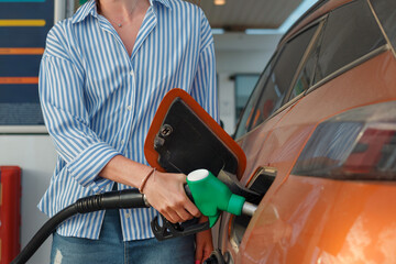A person holds a fuel nozzle, refueling an orange vehicle at a gas station during the afternoon. The atmosphere is busy and practical.