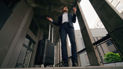Confused business man holding mobile phone in city with suitcase baggage arrive puzzled looking...