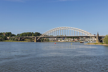 The impressive Waalbrug, a landmark arch bridge, spans the Waal River in Nijmegen under a clear blue sky, connecting the vibrant city with its lush surroundings.