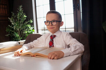 A 7-year-old child in glasses and  dressed school uniform poses at the school table indoor/ The boy learn the book
