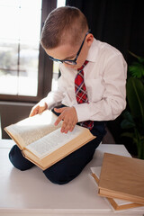 A 7-year-old child in glasses and  dressed school uniform poses at the school table indoor/ The boy learn the book