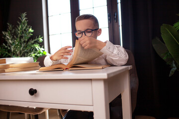 A 7-year-old child in glasses and  dressed school uniform poses at the school table indoor/ The boy learn the book