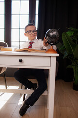 A 7-year-old child in glasses and  dressed school uniform poses at the school table indoor/ The boy learn the book