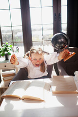 A 7-year-old child in glasses dressed in school uniform poses at the school table indoor. The girl holds globe