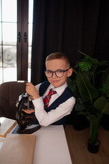 A 7-year-old child in glasses and dressed school uniform poses at the school table indoor. The boy looks to the microscope