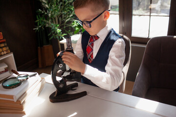 A 7-year-old child in glasses and dressed school uniform poses at the school table indoor. The boy looks to the microscope