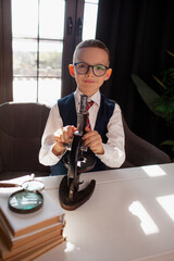 A 7-year-old child in glasses and dressed school uniform poses at the school table indoor. The boy looks to the microscope