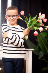 A 7-year-old child in glasses and dresses striped vest, poses at the school table indoor. The boy holds flowers