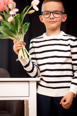 A 7-year-old child in glasses and dresses striped vest, poses at the school table indoor. The boy holds flowers