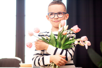 A 7-year-old child in glasses and dresses striped vest, poses at the school table indoor. The boy holds flowers