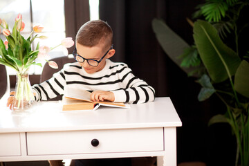 A 7-year-old child in glasses and dresses striped vest, poses at the school table indoor.