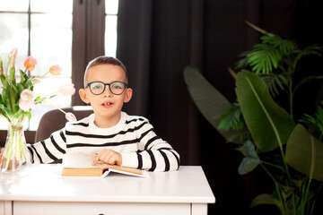 A 7-year-old child in glasses and dresses striped vest, poses at the school table indoor.