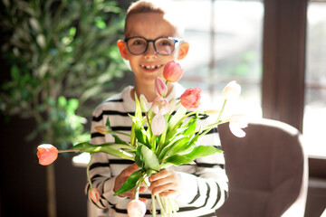 A 7-year-old child in glasses and dresses striped vest, poses at the school table indoor.  The boy holds flowers