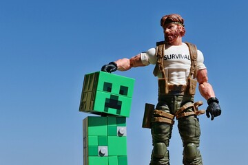 Naklejka premium GI JOE figure of survival instructor Outback, series Classified 2023, stroking head of tamed LEGO Minecraft green explosive mob Creeper on bright daylight sunshine, blue skies in background. 